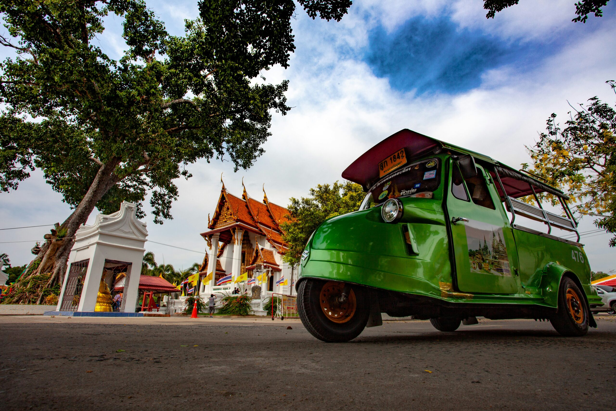 Een karakteristieke groene Thaise tuk-tuk geparkeerd voor een wit tempelcomplex in de straten van Ayutthaya